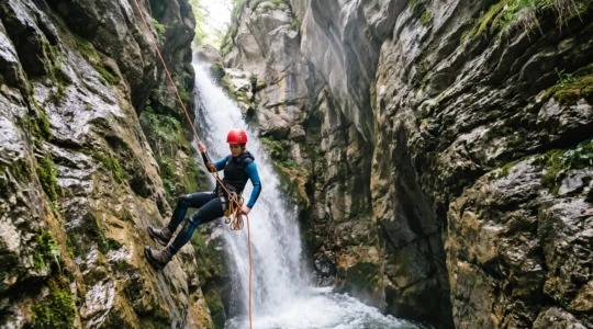 Un pratiquant de canyoning équipé d'un baudrier et d'un casque descend en rappel le long d'une cascade encaissée dans une gorge rocheuse.