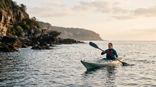 Kayakiste solitaire pagayant calmement le long d'une côte rocheuse au lever du jour, illustrant les débuts sécurisés du kayak de mer